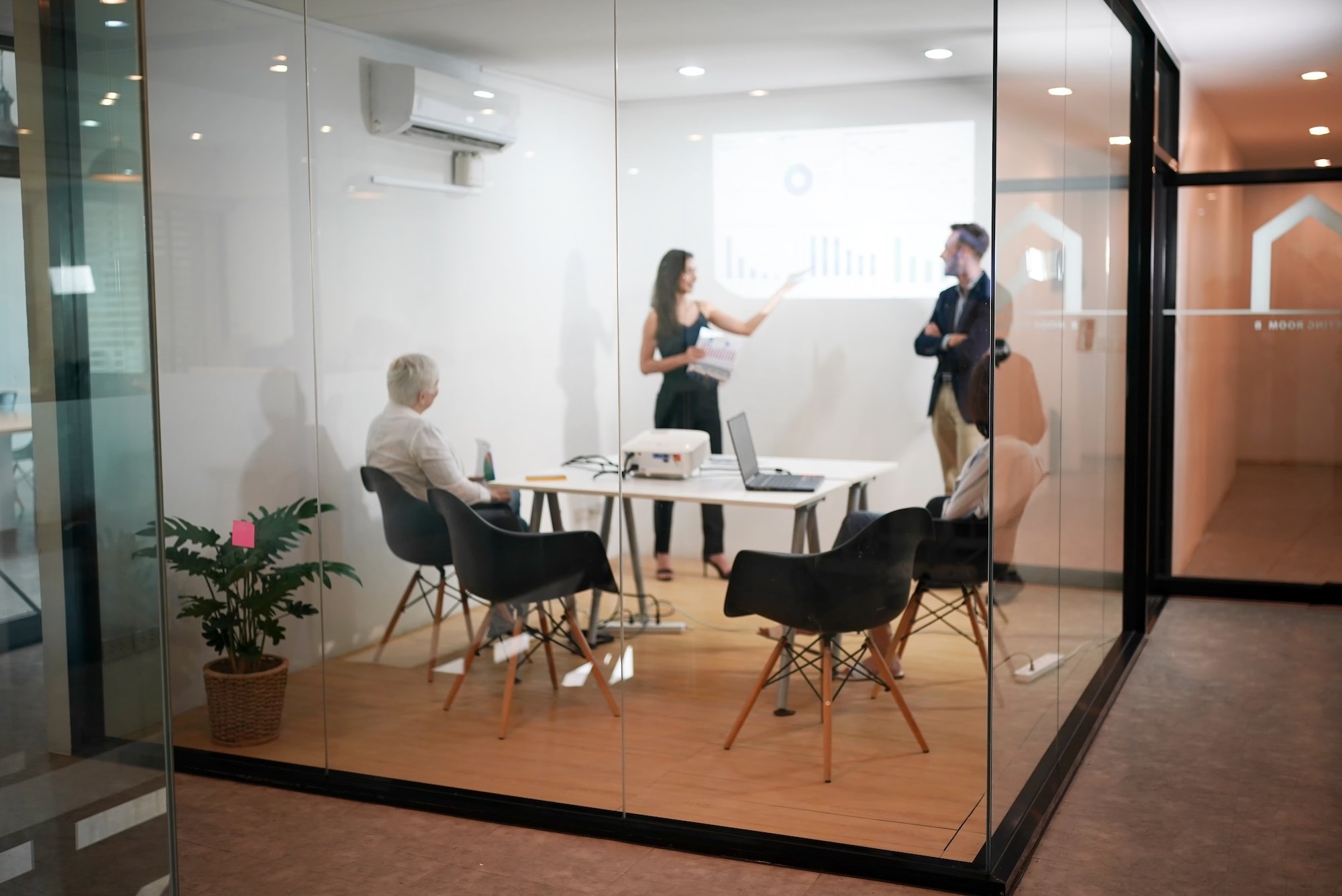A group of people are gathered in a modern, glass-walled office space for a business presentation. The room features a white table with laptops and a projector.